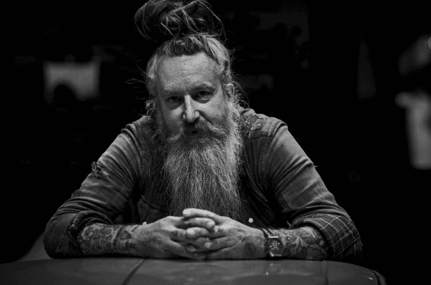 A black and white portrait of a man with a long beard and hair tied in a topknot. He is leaning forward with his hands clasped, wearing a long-sleeve shirt and has tattoos on his arms. The background is dark and out of focus.